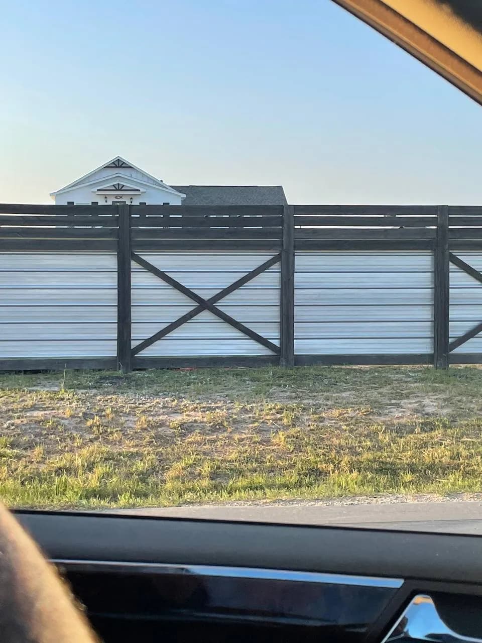 Barda con paneles horizontales de metal corrugado blanco y estructura de madera oscura, con diseños en X. Vista desde calle/exterior.