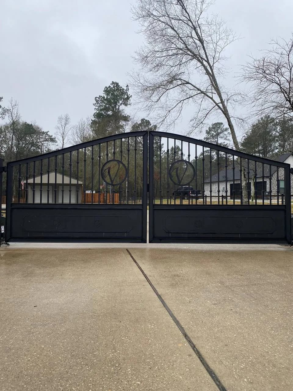 Portón doble de hierro negro con arco superior y círculos decorativos con iniciales (parecen 'O' o 'V'). Base sólida metálica inferior. Camino de concreto.