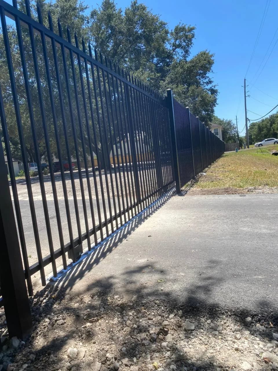 Portón deslizante largo de barras tubulares negras. En riel de piso, divide la calle de un driveway largo hacia una propiedad con muchos árboles.
