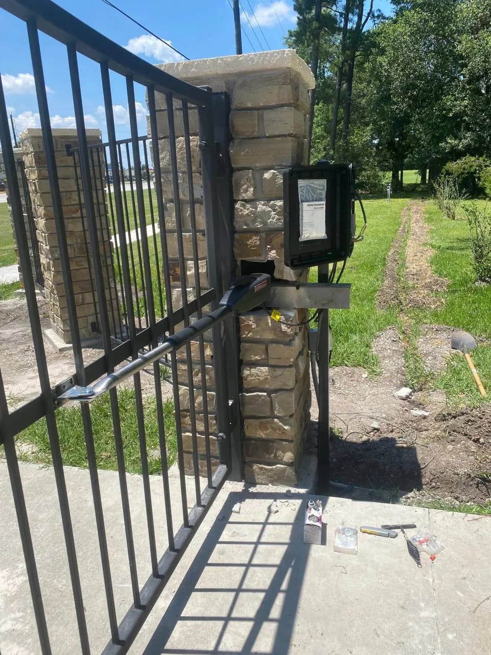 Toma de detalle (close-up) de un brazo motorizado instalado en un portón de hierro forjado, anclado a una columna de piedra caliza/mampostería. Se ve cableado y caja de control abierta.