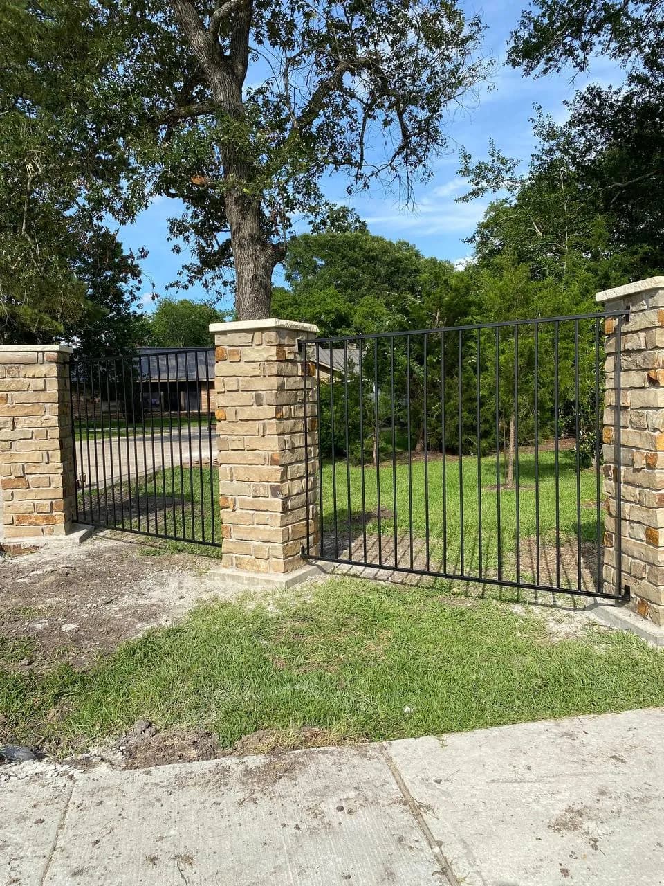 Portón simple de hierro tubular negro entre dos columnas de piedra caliza/mampostería. En medio de un jardín arbolado y pasto verde.