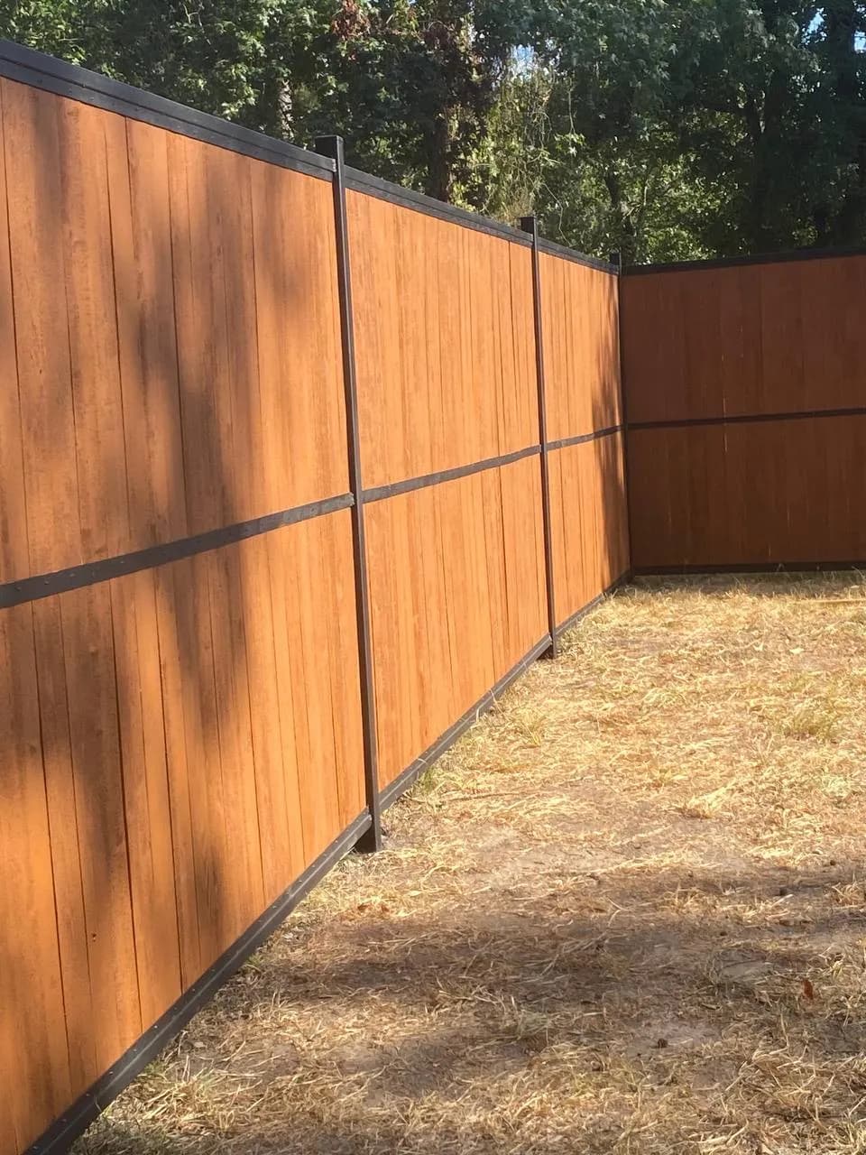 Barda perimetral completamente cerrada. Madera entintada (tono rojizo/cedro) enmarcada y seccionada horizontalmente por rieles de metal negro. Se ven árboles de fondo, luz de día.