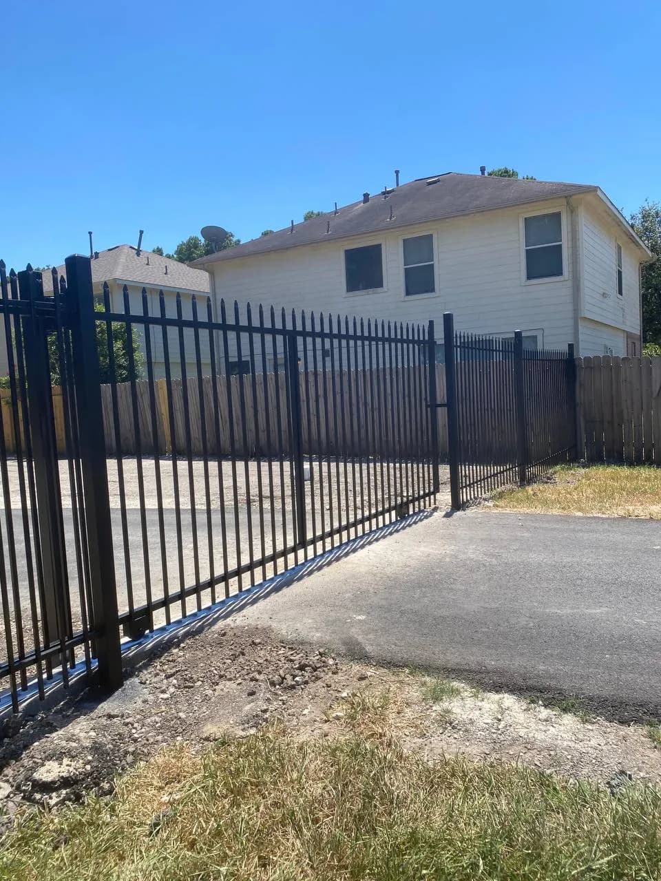 Portón y barda de hierro negro tubular liso separando una propiedad de dos pisos. Portón deslizante con brazo motorizado negro visible abajo.