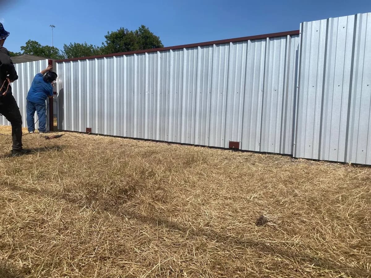 Barda perimetral muy larga de metal corrugado galvanizado/plateado con marco superior y bases en color vino/rojo. Dos trabajadores instalando/soldando.