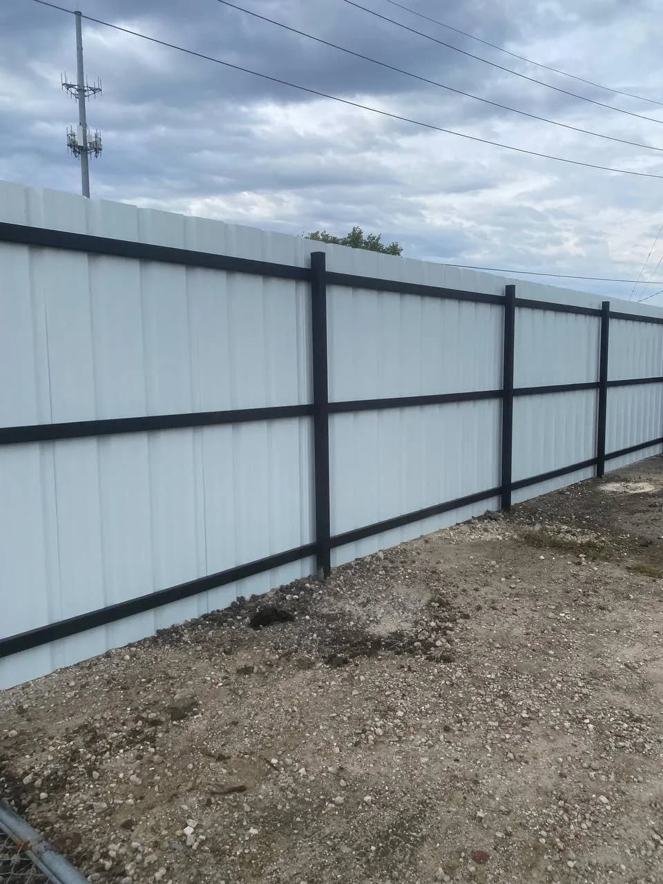 Barda de metal corrugado blanco con postes y marcos transversales (3 horizontales) en hierro negro, bordeando un terreno de grava. Toma exterior bajo nubes.