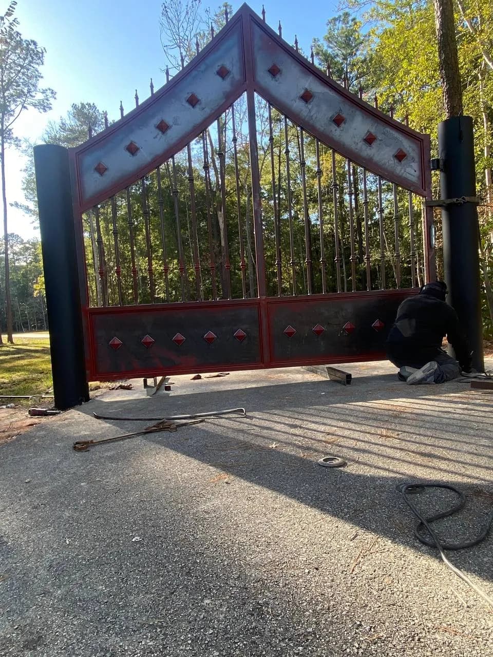 Trabajador instalando un portón asimétrico espectacular en forma de 'A' (pico), con rombos en rojo/vino y paneles metálicos opacos. Diseño muy robusto para entrada de rancho.