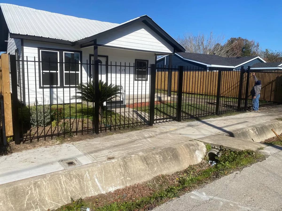 Barda frontal de hierro tubular negro simple frente a una casa blanca (con número 117). Un instalador trabajando en la esquina. Divide banqueta de concreto de jardín.
