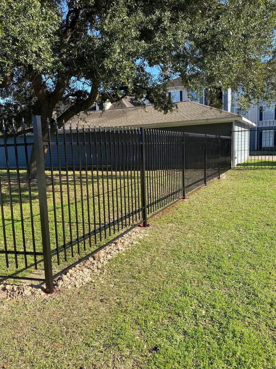 Barda de hierro negro con puntas ornamentales (fleur-de-lis o lanzas) rodeando una propiedad residencial con pasto. Postes cuadrados negros.