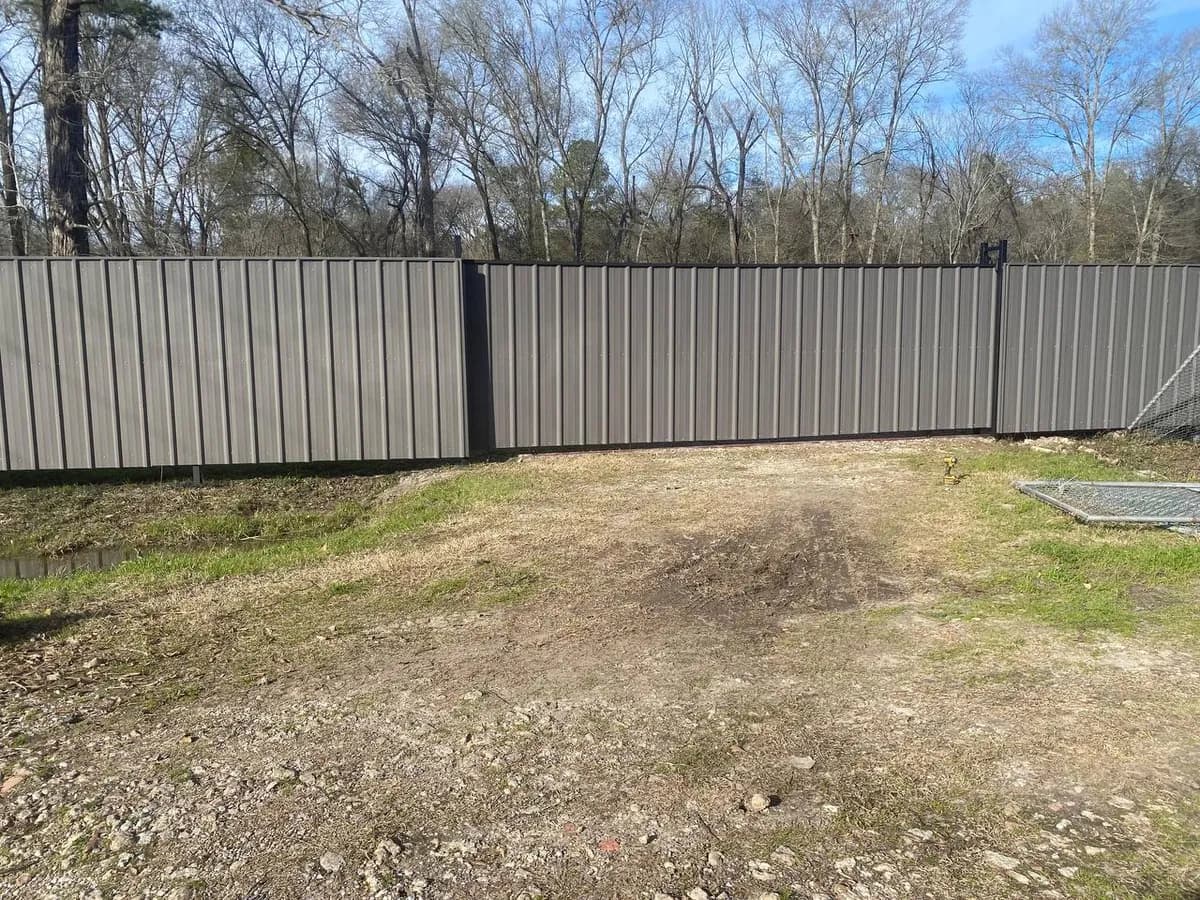 Barda opaca y alta hecha de metal corrugado pintado en gris oscuro/café con marco negro. Ubicada en zona boscosa. Similar al primer video.