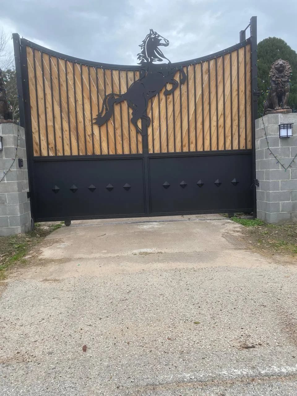 Portón masivo impresionante con un caballo rampante (silueta en metal negro) arriba en el centro, relleno de varas/tablas de madera clara en diagonal, base metálica sólida con rombos, y dos estatuas de leones en pilares de bloque a los lados.