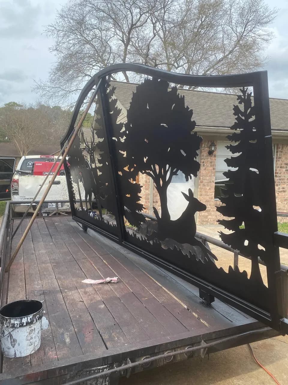 Toma del portón subido a una plataforma/remolque antes o después de la instalación. Diseño asombroso de arte en metal cortado a láser (plasma): escena de bosque con un árbol central, un ciervo parado a la izquierda y un ciervo acostado a la derecha.
