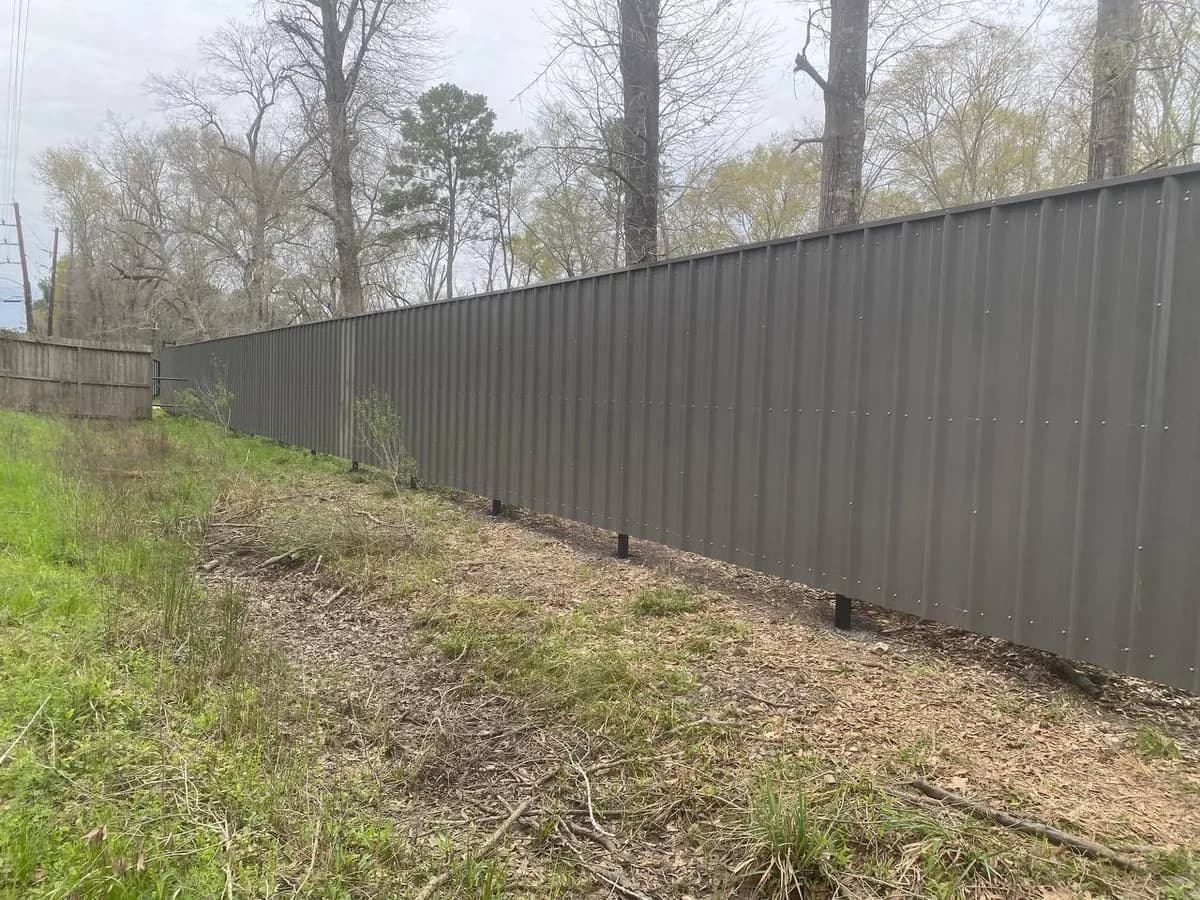 Otra perspectiva de la barda alta gris oscura/café de paneles corrugados verticales y marco negro bordeando el bosque, vista exterior en declive hacia pasto.