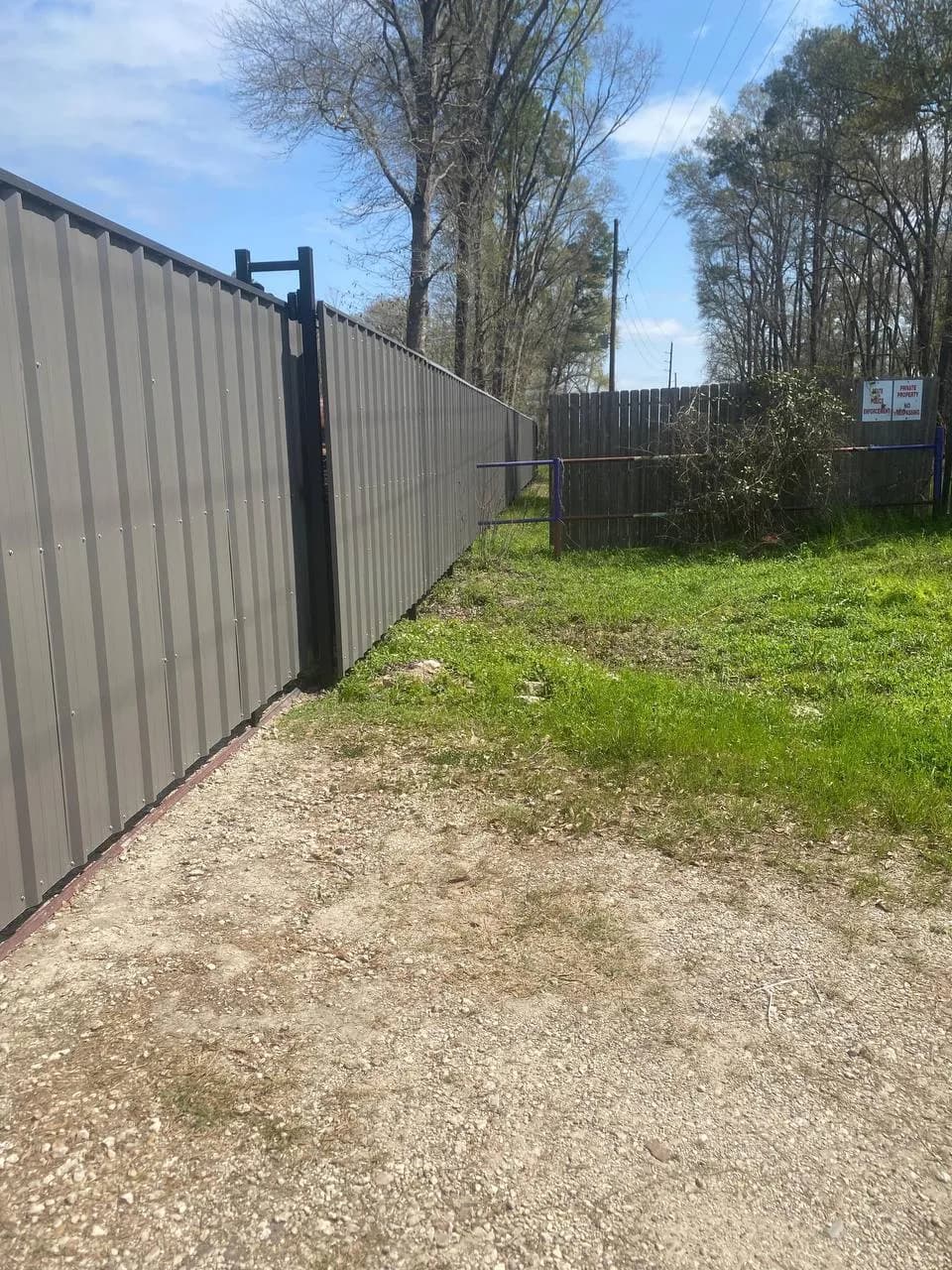 Barda gris oscura (la misma de fotos anteriores) junto a una valla de malla ciclónica vieja con letreros de 'Private Property'. Muestra el contraste del antes (malla) y el después (corrugado opaco).