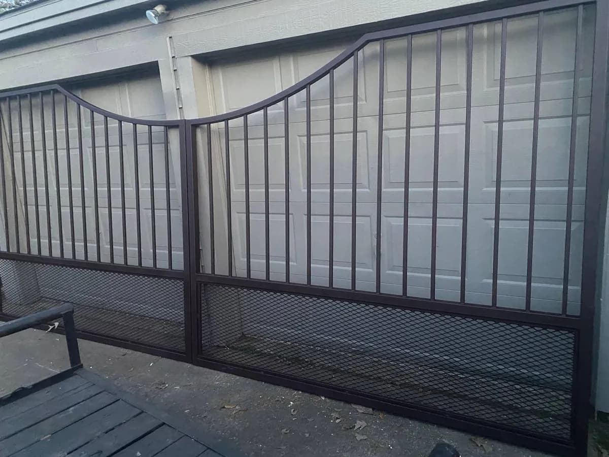 Portón doble asimétrico (lado izquierdo cae en curva). Hierro tubular superior y malla de metal expandido en el tercio inferior. Recién fabricado, apoyado frente a un garage.