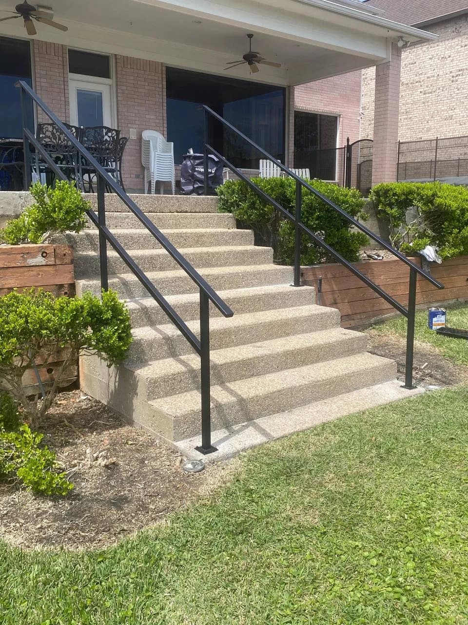 Pasamanos (Railing) instalado en las escaleras exteriores de concreto del patio trasero de una casa de ladrillo. Hierro negro, diseño limpio. (Ojo: Nueva subcategoría identificada).