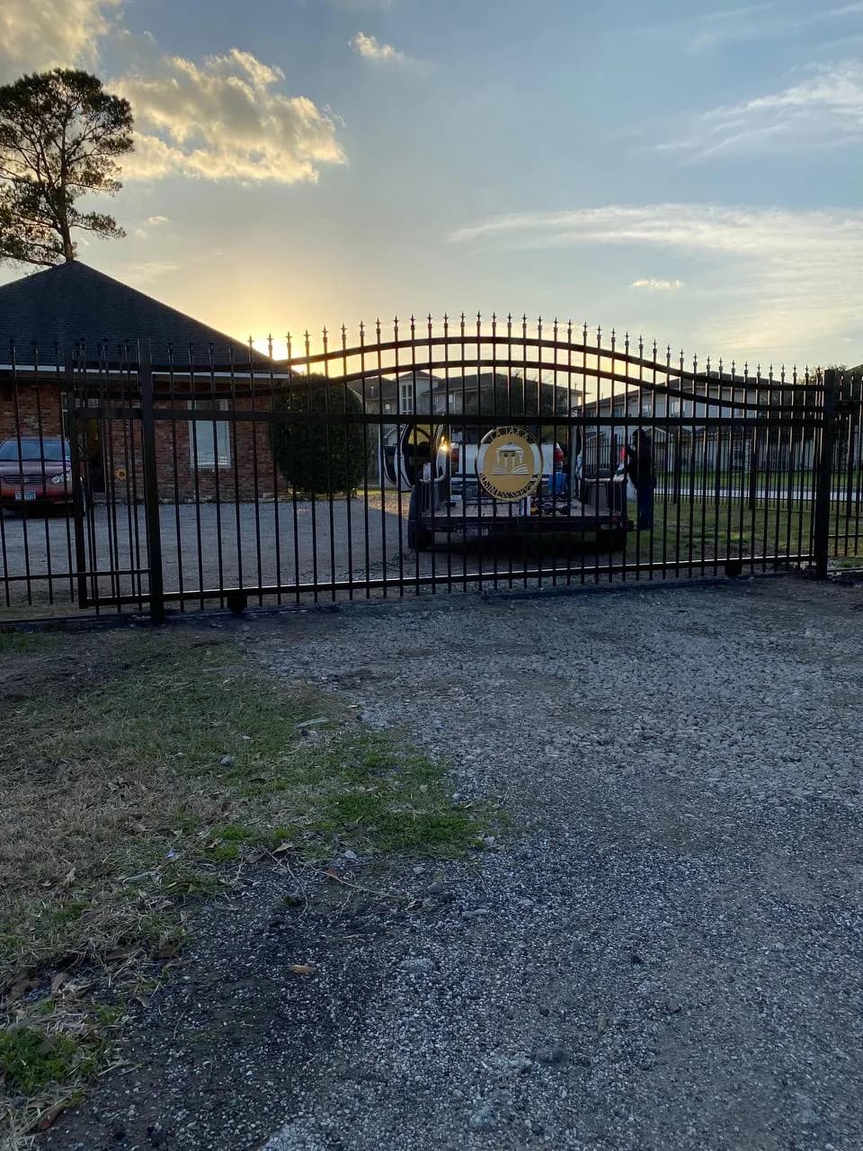 Portón largo deslizante (o doble) de hierro negro, con arco en la parte superior y escudo decorativo central (dorado/amarillo) con letras 'V' y diseño arquitectónico. Vista de atardecer.