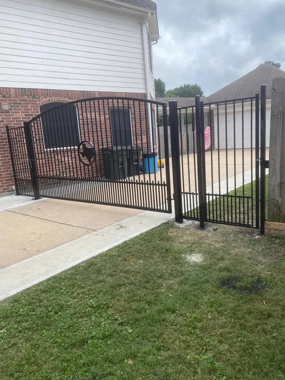 Portón con arco asimétrico superior (slider) y puerta peatonal integrada o adyacente. Diseño en hierro negro con un escudo central con la forma del estado de Texas y una estrella en medio.