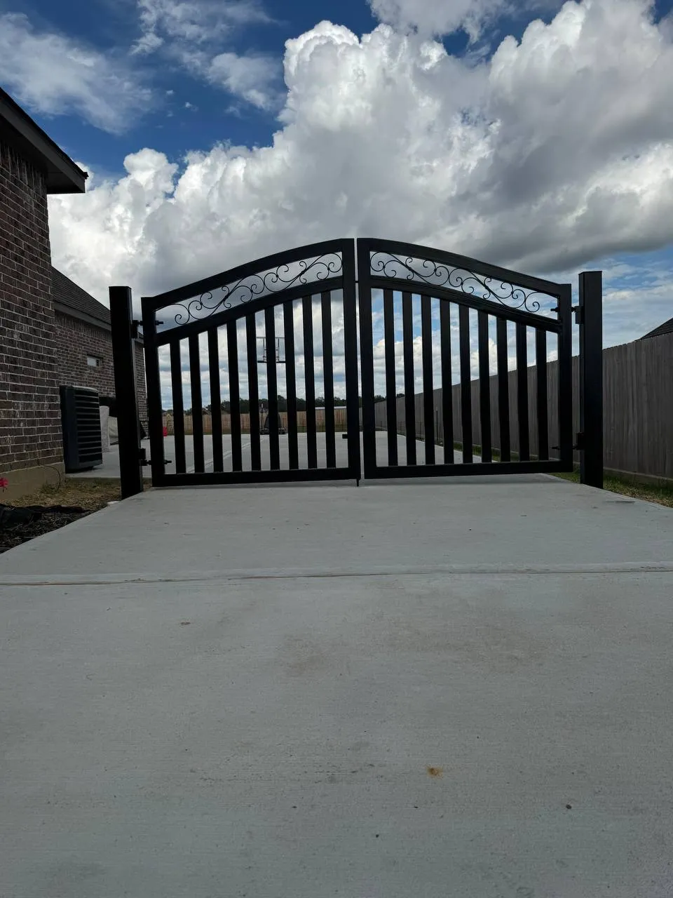 Portón doble elegante con arco pronunciado en hierro negro. La parte superior tiene diseños curvos (scrolls) muy detallados. El driveway es de concreto.