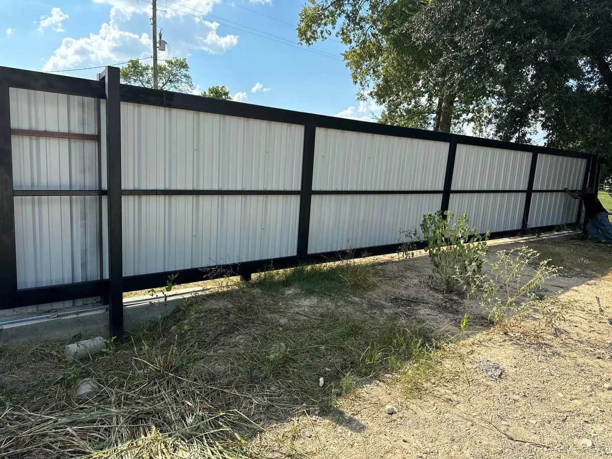 Barda perimetral muy larga de lámina corrugada blanca/clara con postes y marco negro/oscuro. Construida sobre una base/guarnición de concreto.