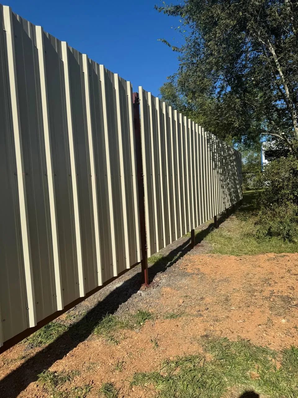 Toma posterior de la barda corrugada blanca (file_33). Muestra los postes redondos tubulares oscuros enterrados/sujetando la estructura. Vista de campo con árboles de fondo.