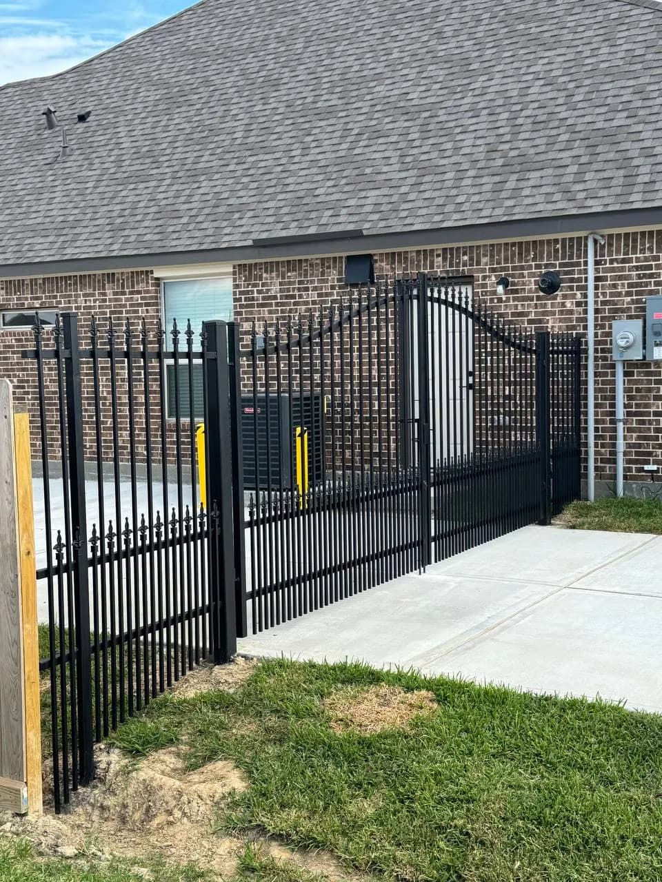 Sistema completo de portón doble deslizante y barda a juego. Hierro negro con puntas ornamentales (fleur-de-lis). Arco sutil en el portón. Protege el lateral de una casa de ladrillo rojo.