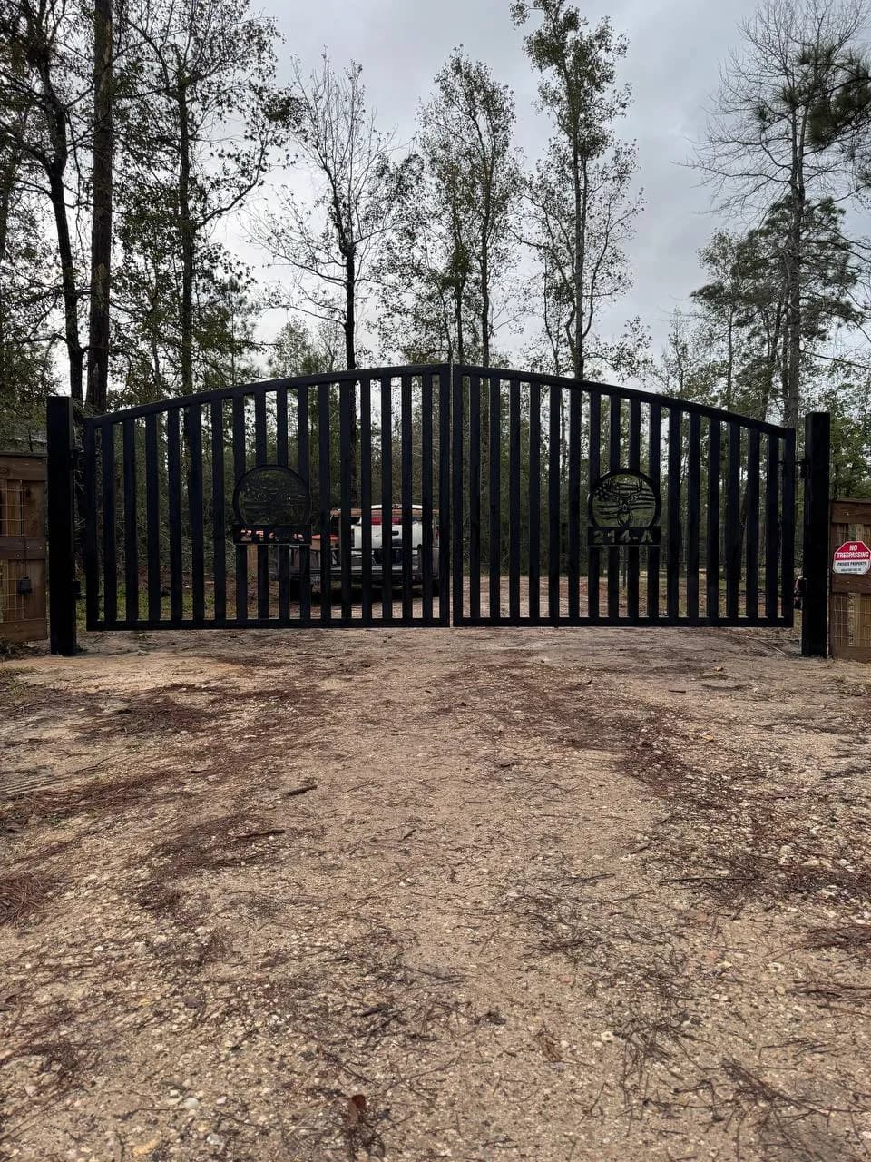 Portón doble imponente en medio del bosque. Hierro negro, arco superior y diseños circulares con escudos de águila y la bandera de EE.UU., junto con los números '214-A'. Letrero de 'No Trespassing'.