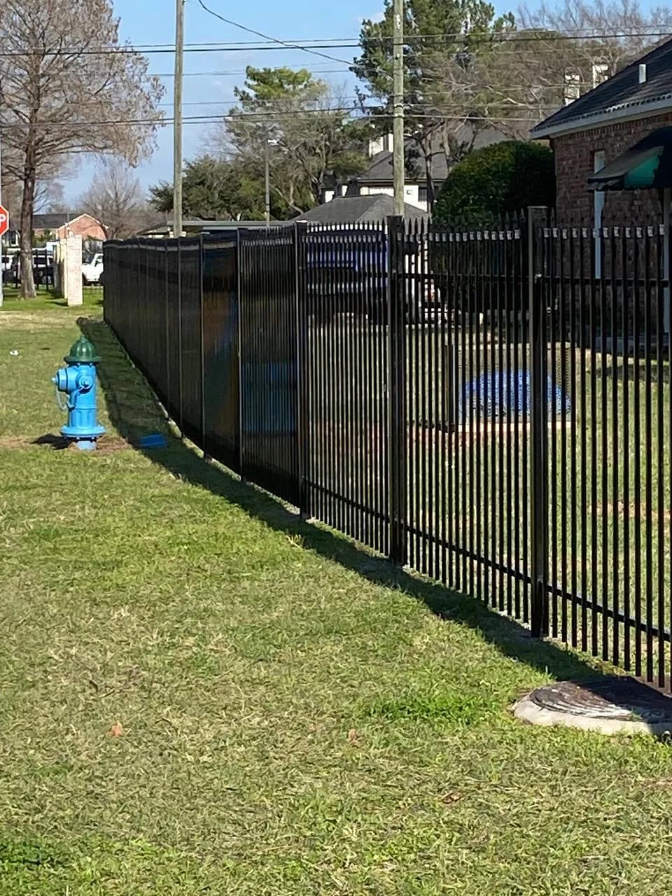Barda tubular negra con puntas decorativas en la parte superior, bordeando el frente de una casa de ladrillo. Toma de día soleado.