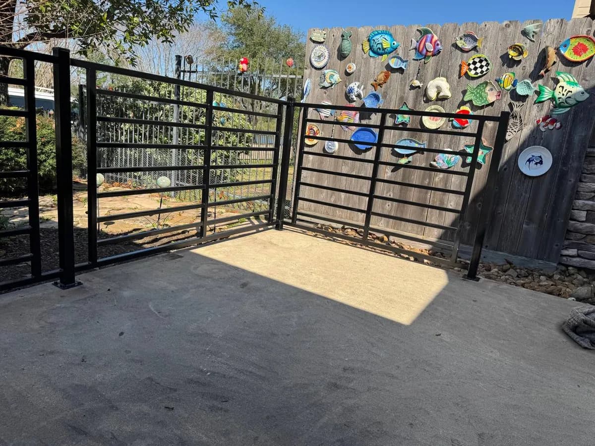 Barda perimetral pequeña/cercado en patio trasero. Estructura de hierro negro con líneas horizontales gruesas enmarcadas, cubriendo parte de una valla de madera trasera con decoraciones de platos.