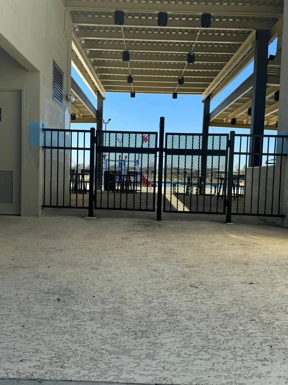 Portón doble pequeño bajo una cubierta industrial. Hierro negro con relleno de malla metálica expandida. Da acceso a lo que parece un área de alberca o parque acuático comercial.