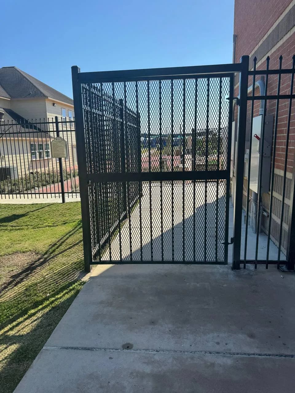 Portón peatonal y barda lateral del mismo complejo de la foto 42 (alberca). Estructura negra con malla expandida, junto a un muro de ladrillo e instalaciones eléctricas.