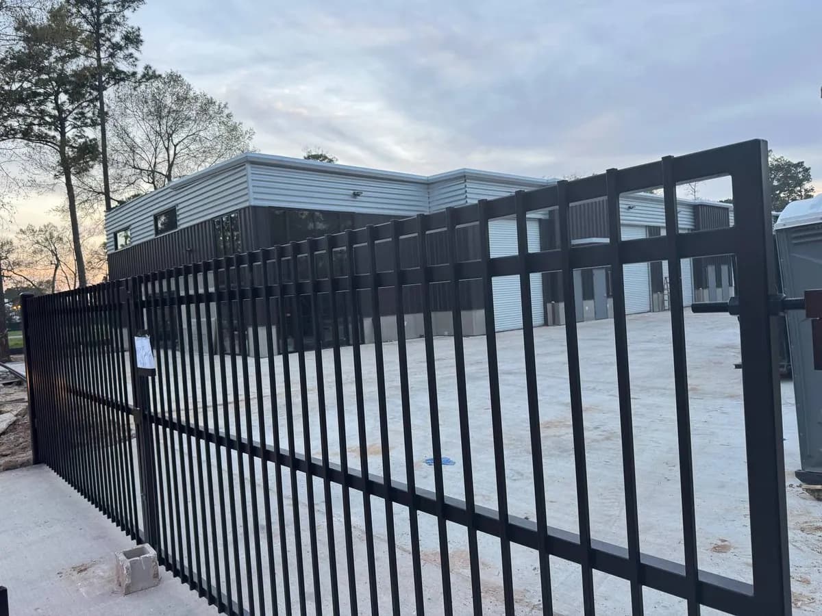 Larga barda de hierro negro estilo 'flat top' (sin puntas, borde liso superior) protegiendo un estacionamiento de concreto y la fachada de una nave industrial comercial de acero gris.