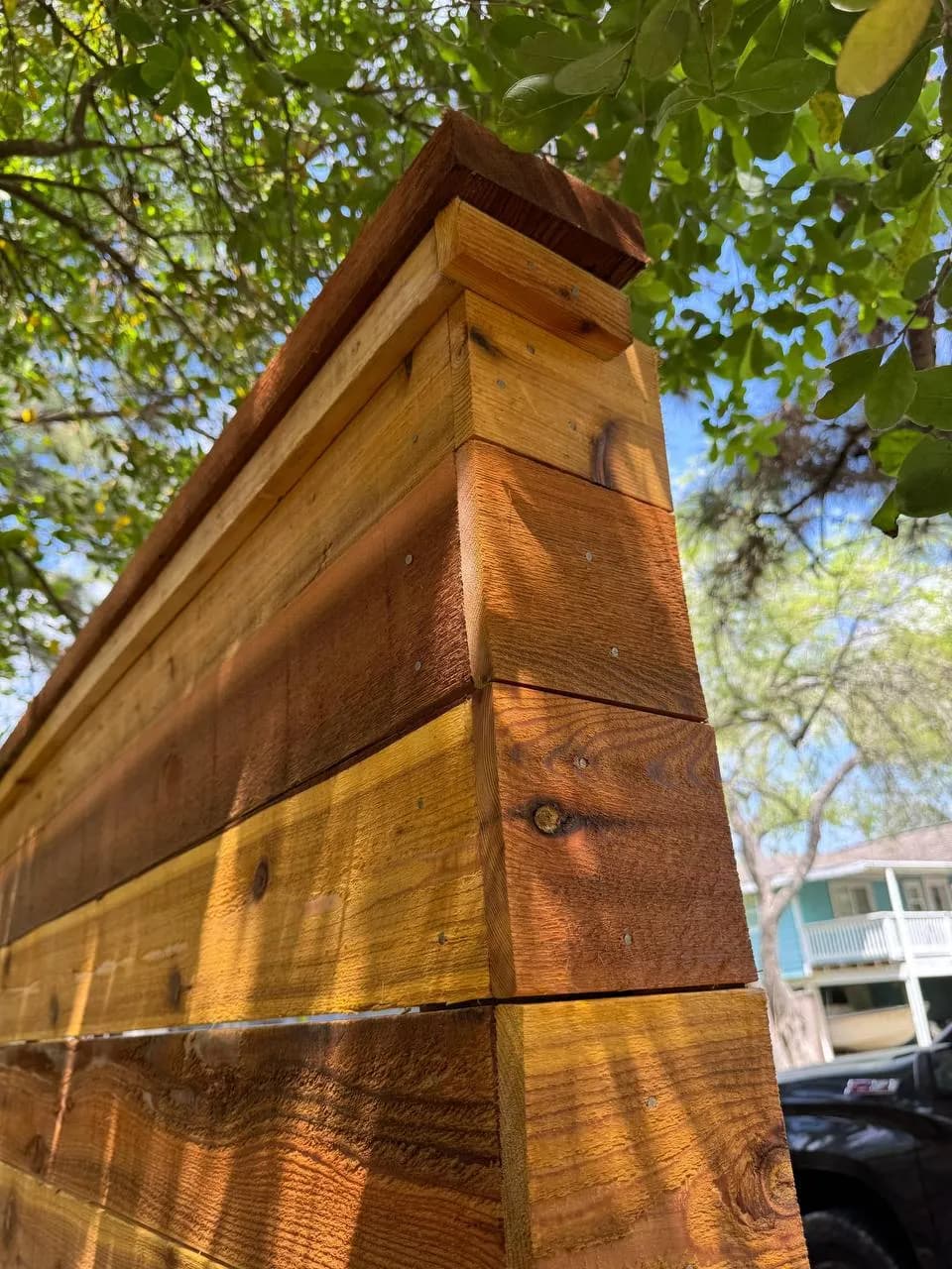 Close-up a la esquina superior de una barda de madera con acabado natural claro (pino/cedro). Las tablas están horizontales con marco grueso superior. Trabajo muy limpio de carpintería.