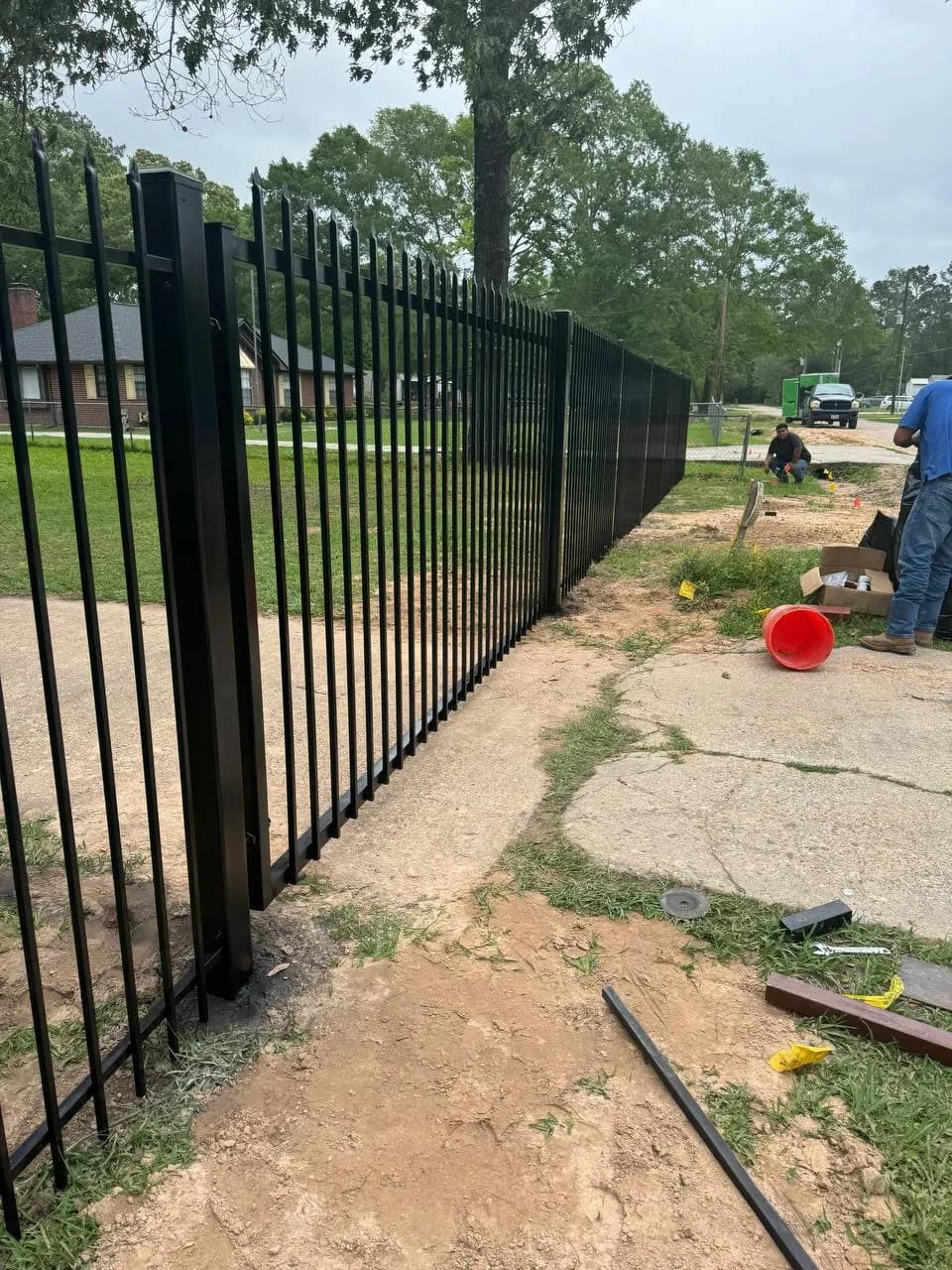 Larga barda de hierro tubular negro con puntas ornamentales altas (spear tops) a lo largo de un driveway. Se ven dos instaladores trabajando en el pasto ajustando postes.