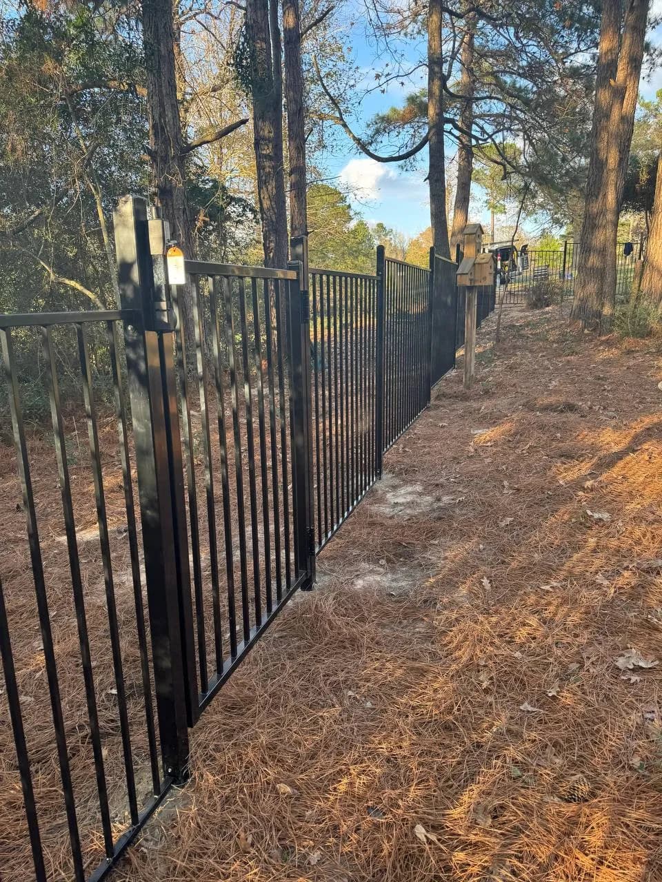 Barda perimetral en área boscosa/con pendiente de pinochas. Incluye una puerta peatonal sencilla. Estilo flat top con barras simples.