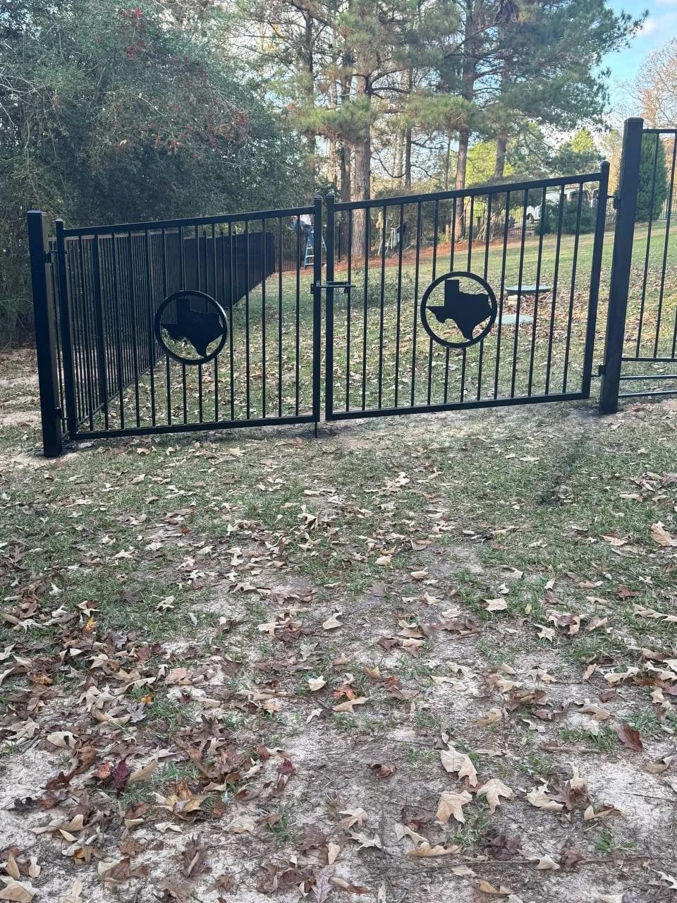 Pequeño portón doble de herrería negra (estilo flat top) cruzando un jardín/patio lateral con hojas secas. Tiene círculos centrales con el estado de Texas calado adentro.