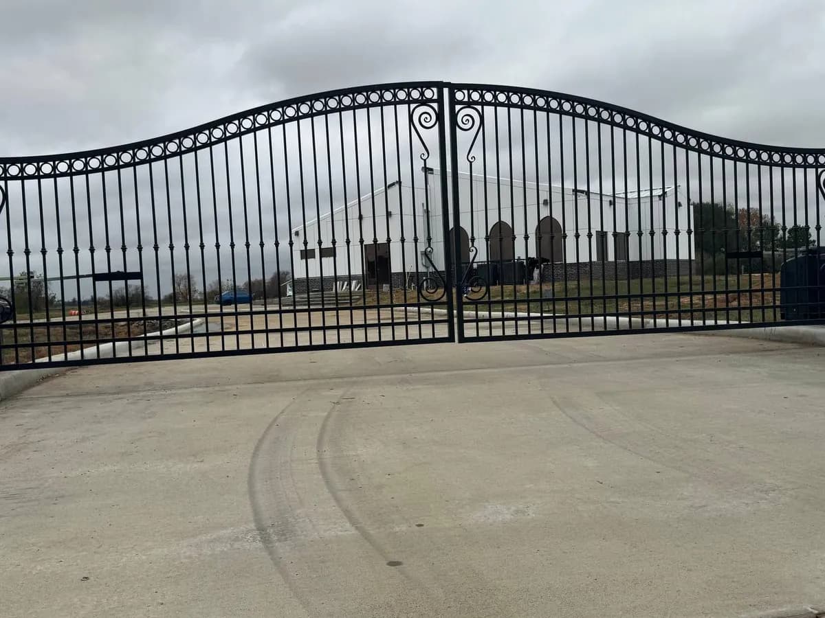 Enorme portón doble (muy ancho) de hierro tubular negro con un arco ondulado superior. Diseños curvos (scrolls) en los postes centrales. En el fondo se ve un edificio blanco grande que parece iglesia o salón de eventos.