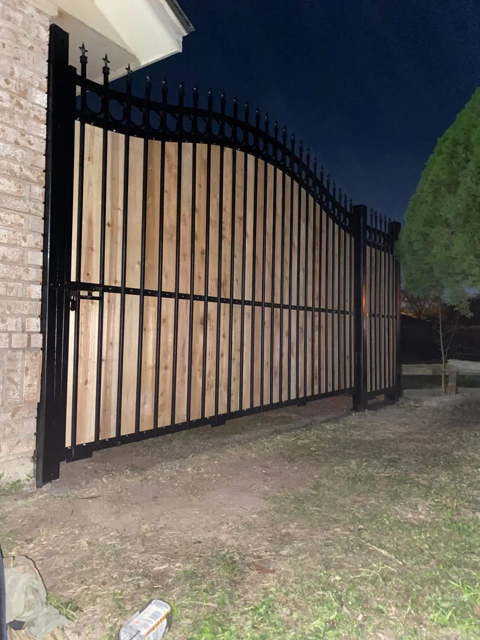 Portón doble de hierro forjado negro (arco superior con puntas) con paneles verticales de madera clara (cedro o similar) rellenando la estructura. Da a pared de ladrillo.