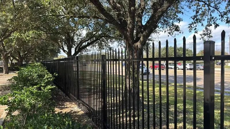 Extensa barda de herrería tubular negra con puntas anchas en la parte superior. Bordeando una vialidad o estacionamiento grande con árboles y césped.