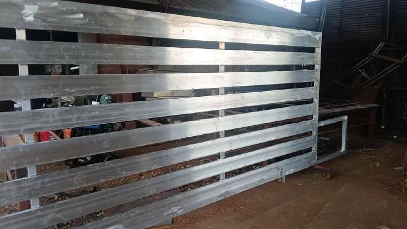Foto dentro del taller de herrería (welding shop). Se ve la estructura de un portón (o barda) de barras planas anchas y horizontales en metal crudo plateado brillante (aluminio o acero sin recubrir), sobre mesas de trabajo.