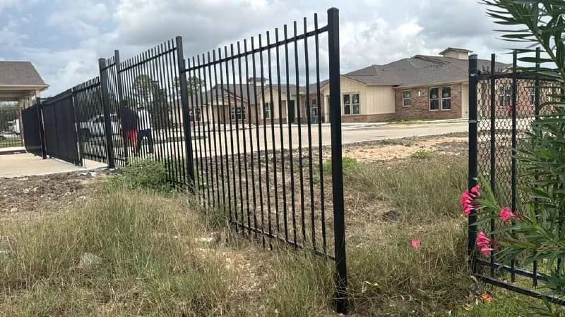 Barda perimetral negra de herrería estándar, colocada sobre pasto irregular, protegiendo un complejo nuevo de viviendas/departamentos de ladrillo. Ojo: a la derecha se ve cómo conecta o reemplaza una cerca vieja de malla ciclónica.