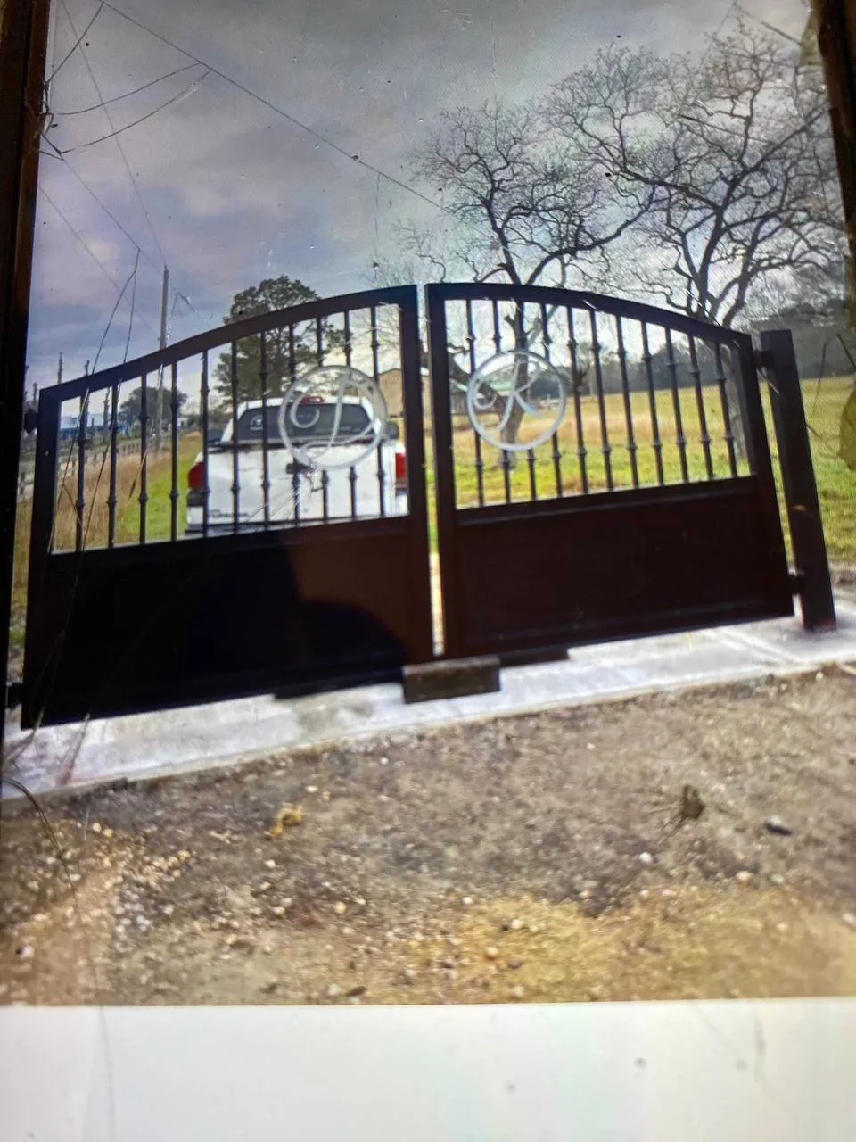 Portón doble de hierro negro, arco superior, placa metálica ciega abajo, círculos en el centro con iniciales cursivas ('C' y 'K' o similar). Toma desde una pantalla/monitor.