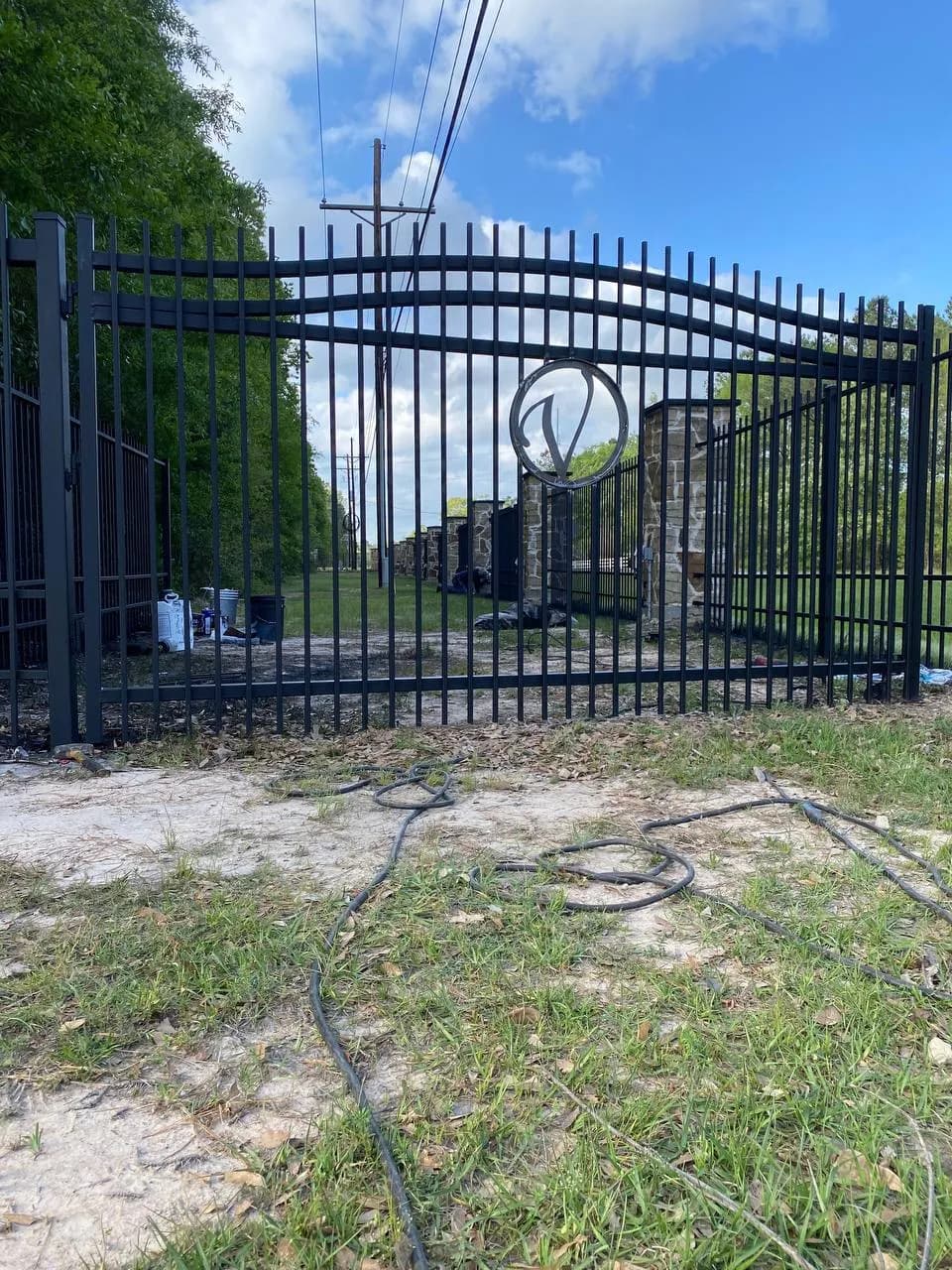 Portón doble largo de hierro negro con arco suave, círculo decorativo central (parece una 'V'). Se sostiene en columnas de mampostería (piedra). Se ve herramienta y cables tirados, parece recién instalado.