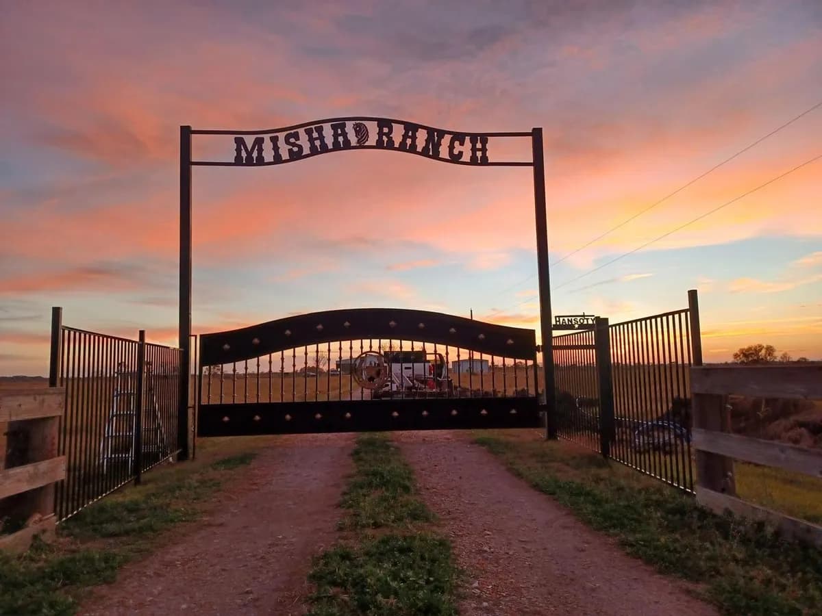 Gran arco de entrada 'MISHAD RANCH' con portón de hierro forjado bajo un cielo de atardecer.