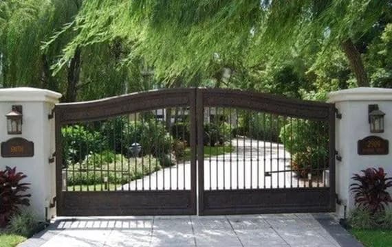 Portón doble ornamentado con acabado en tono bronce/cobre, columnas blancas y faroles clásicos.